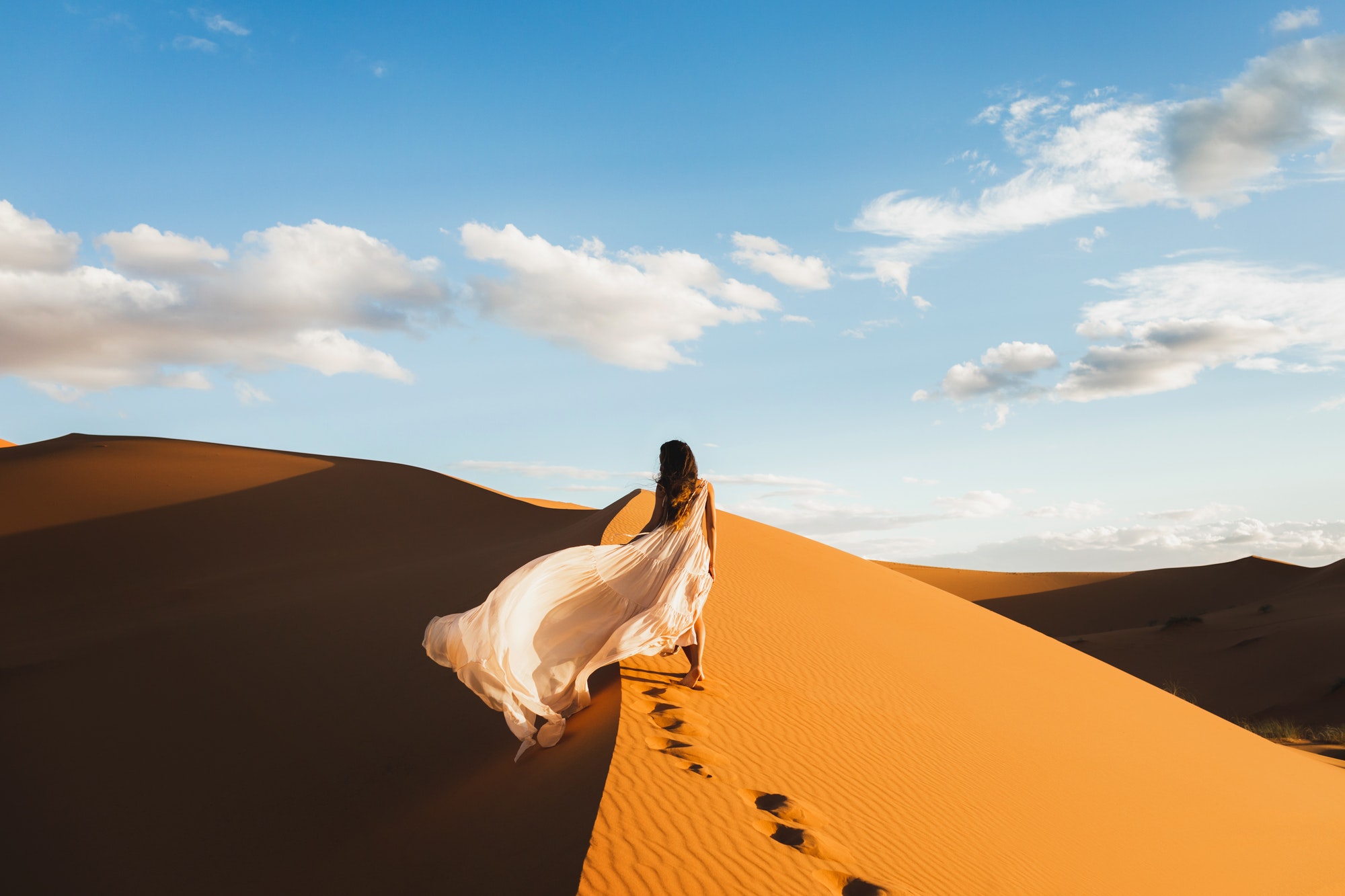 Woman in amazing silk wedding dress with fantastic view of Sahara desert sand dunes in sunset light